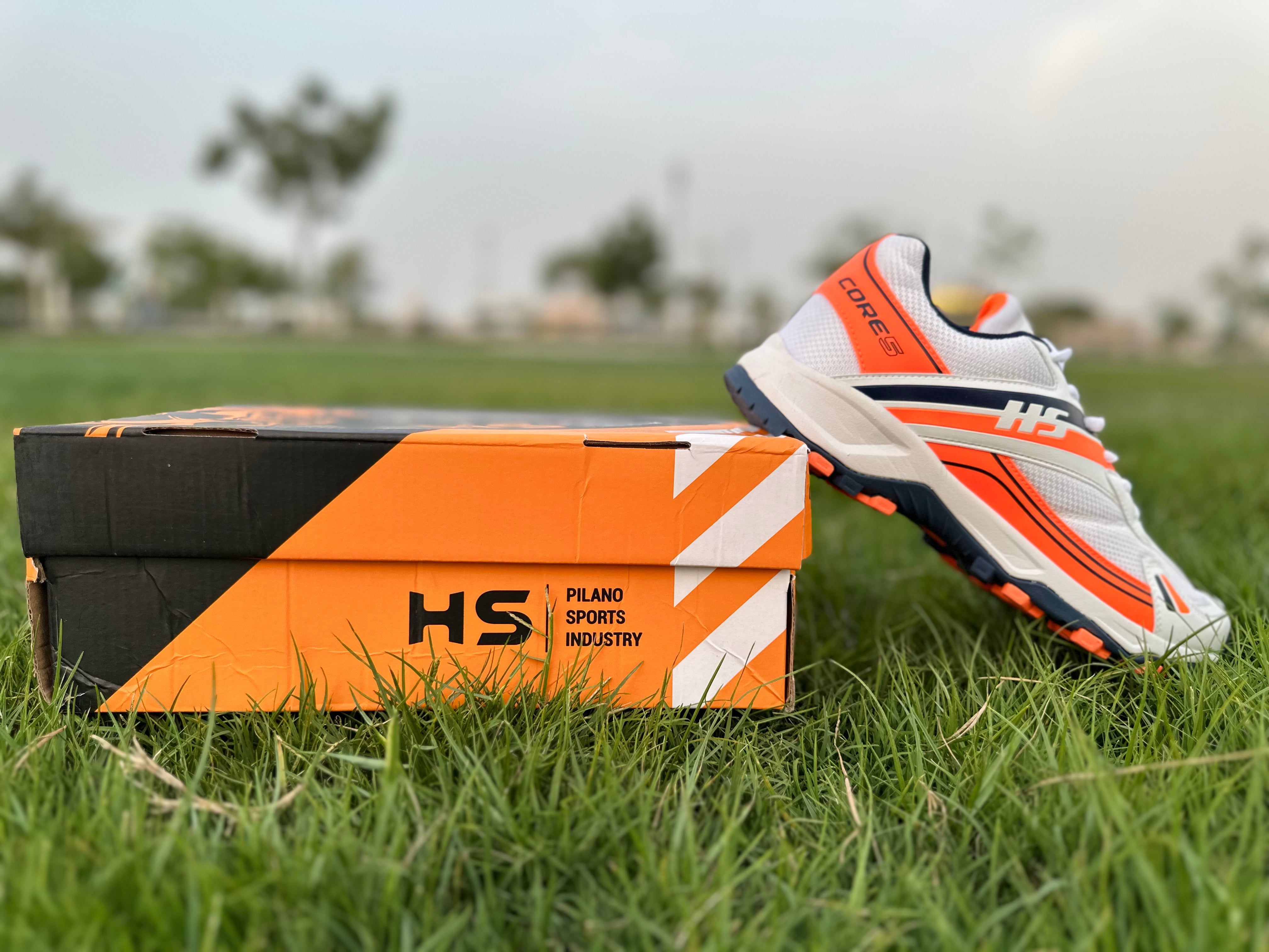 HS Cricket Shoes-Core-5 in orange color beside packaging on grass, lightweight cricket shoes for performance, EUR 42.